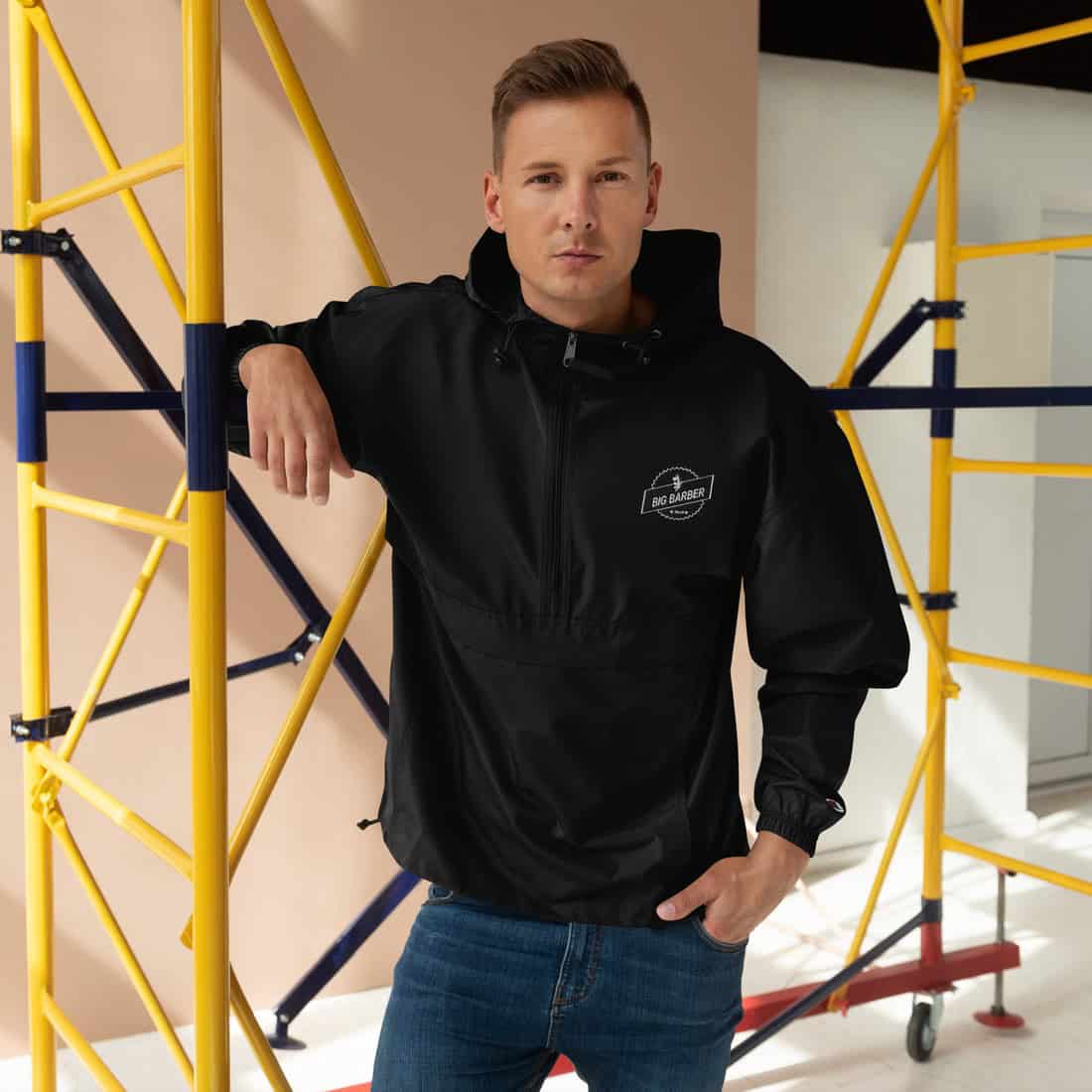 Stylish young man in black Big Barber Deluxe jacket leaning against yellow scaffold at trendy barber shop. Haircut and grooming fashion focus, modern men's barbering, confident style, professional grooming services.